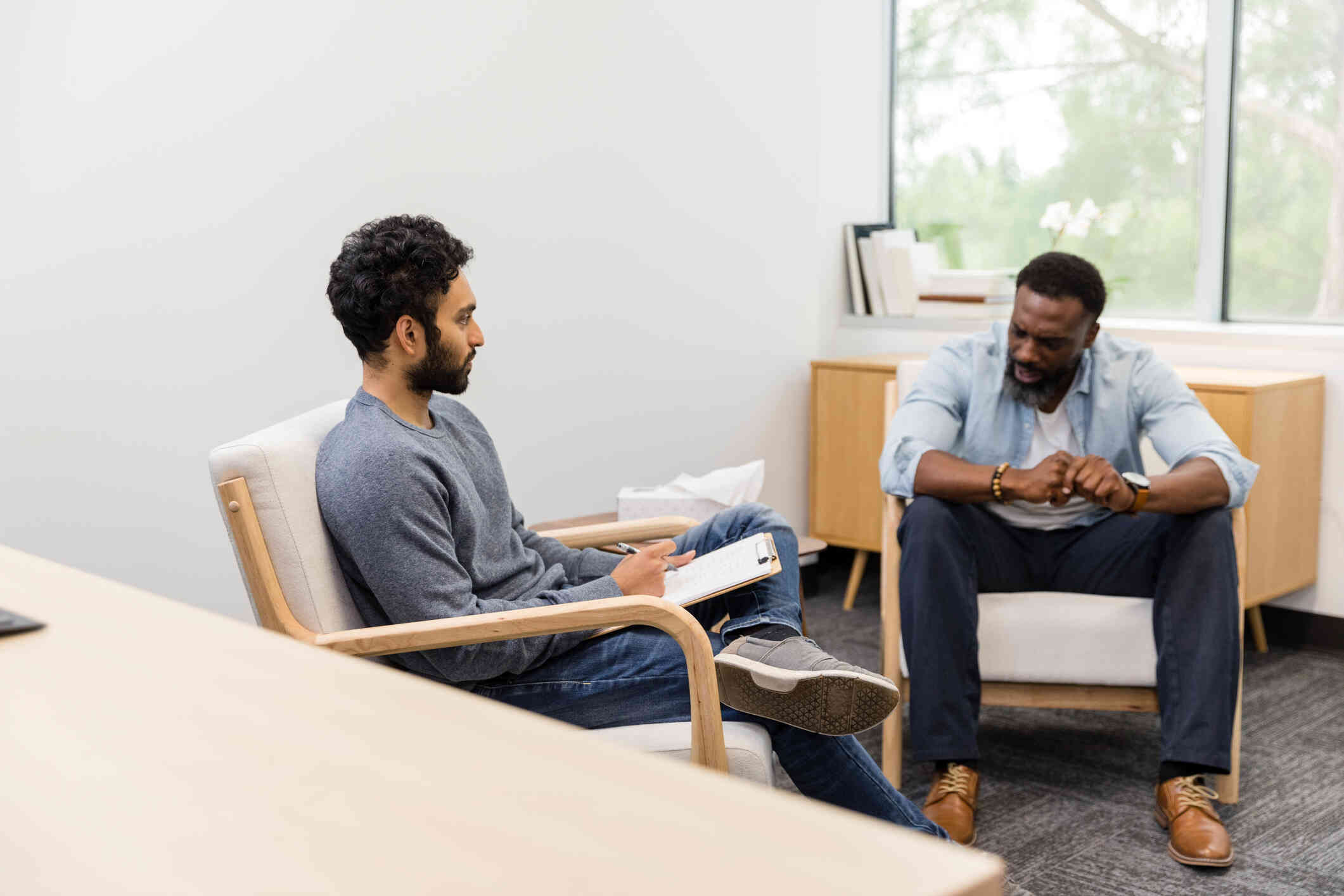 A middle aged man looks distressed as he sits huncehd over in a chair and talks to the male therapist sitting across from hm during a therapy session.